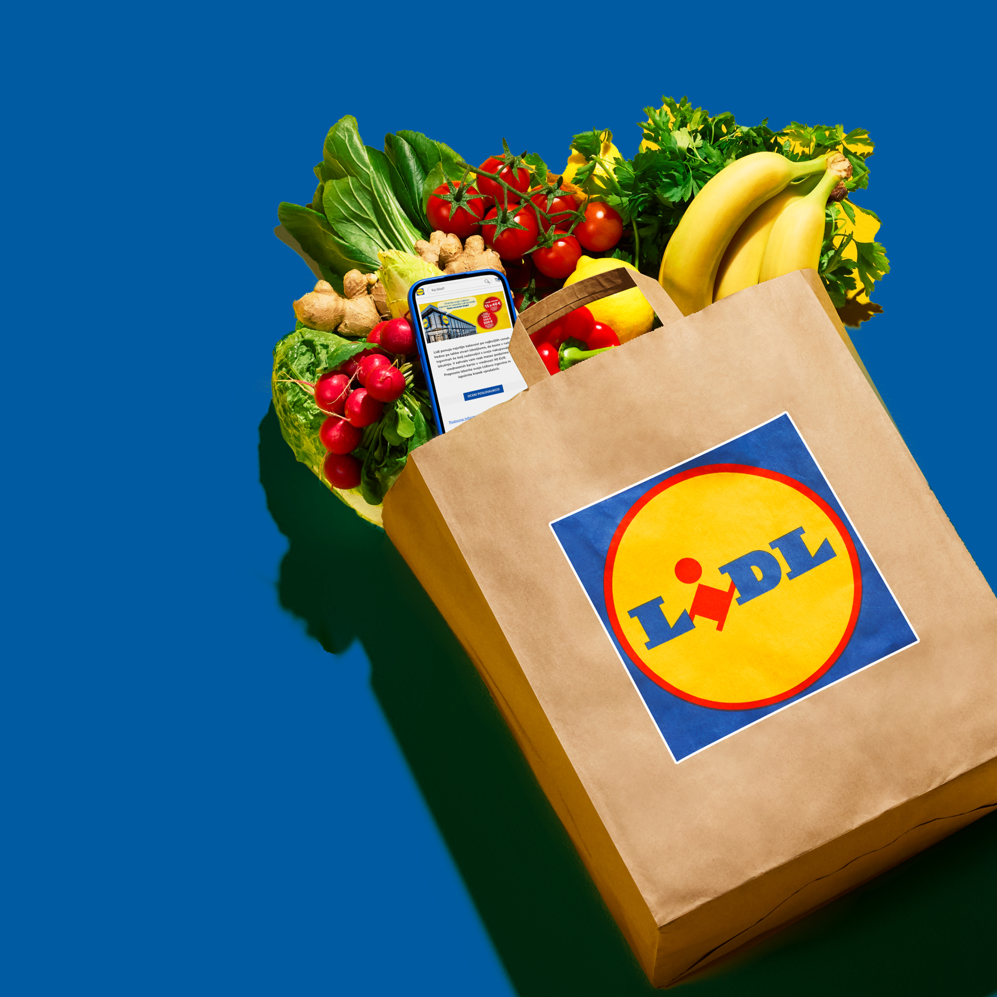 Nakupovalna vrečka Lidl, polna sveže zelenjave in sadja, s telefonom, ki prikazuje aplikacijo Lidl.