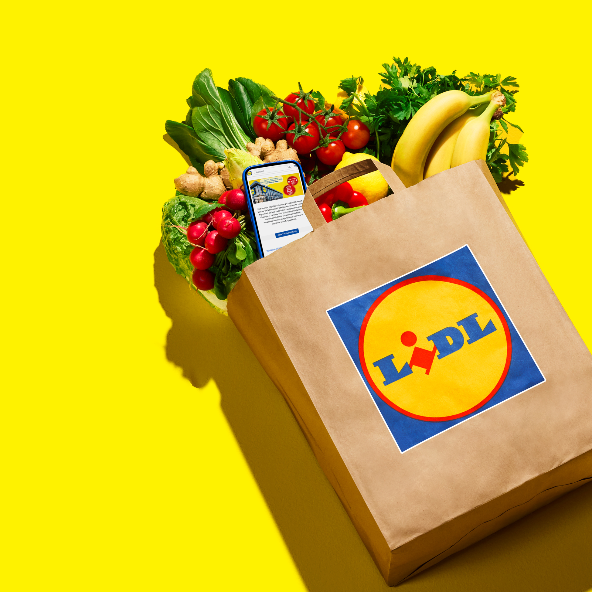 Nakupovalna vrečka Lidl, polna sveže zelenjave in sadja, s telefonom, ki prikazuje aplikacijo Lidl.