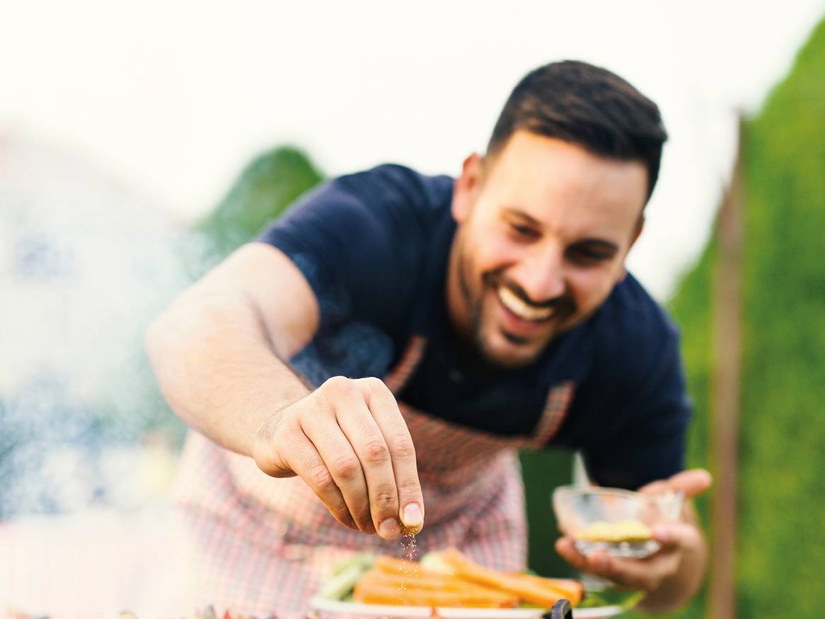 Nasmejan moški posipa začimbe po zelenjavi na žaru.
