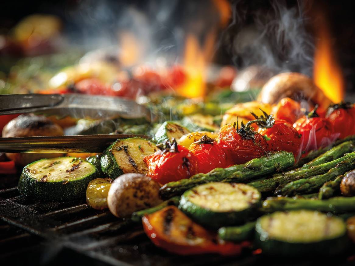 Zelenjava na žaru, vključno z bučkami, paradižnikom in šparglji, s plameni in dimom.