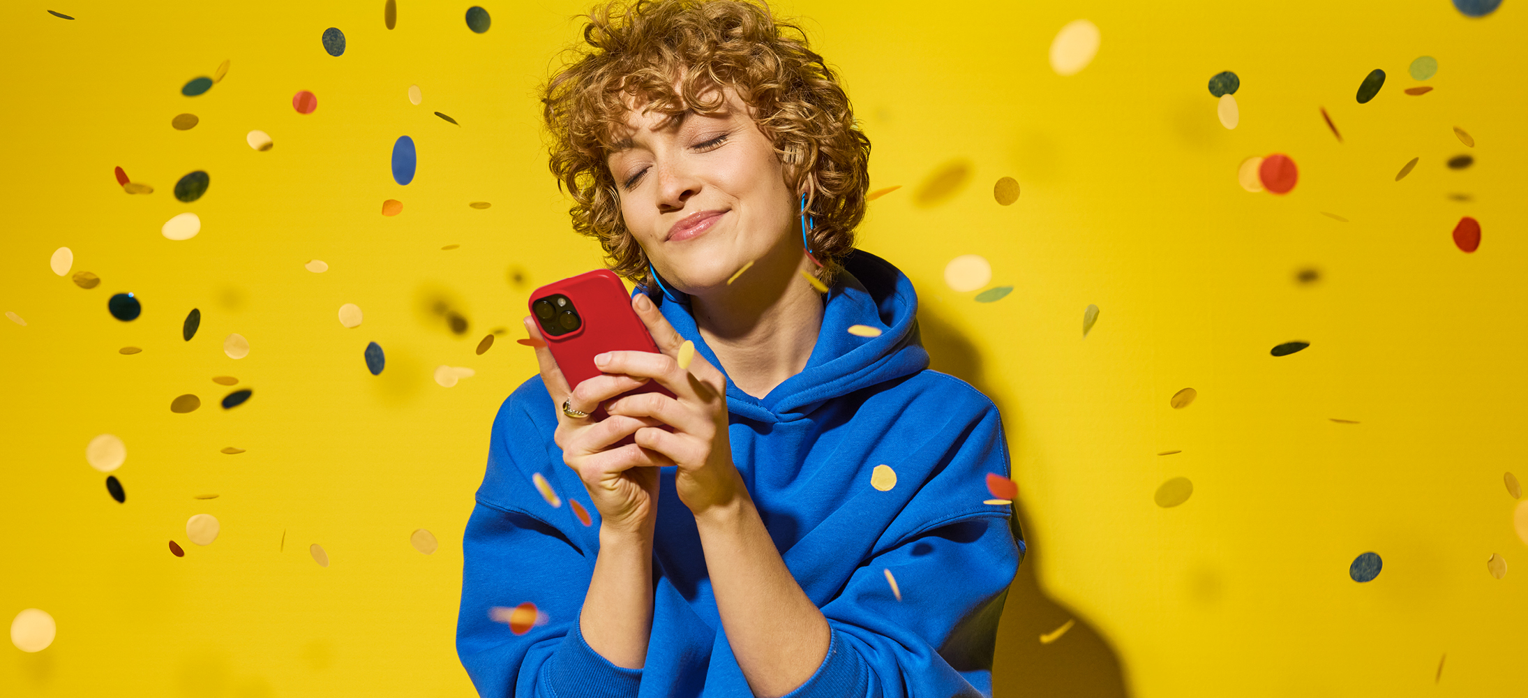 Ženska v modri jopici drži rdeč telefon, medtem ko okoli nje letijo konfeti.