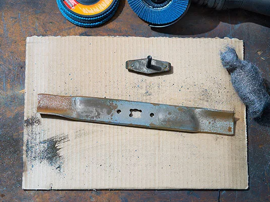 Zarjavela rezila kosilnice na kartonu z brusnimi ploščami in jekleno volno.