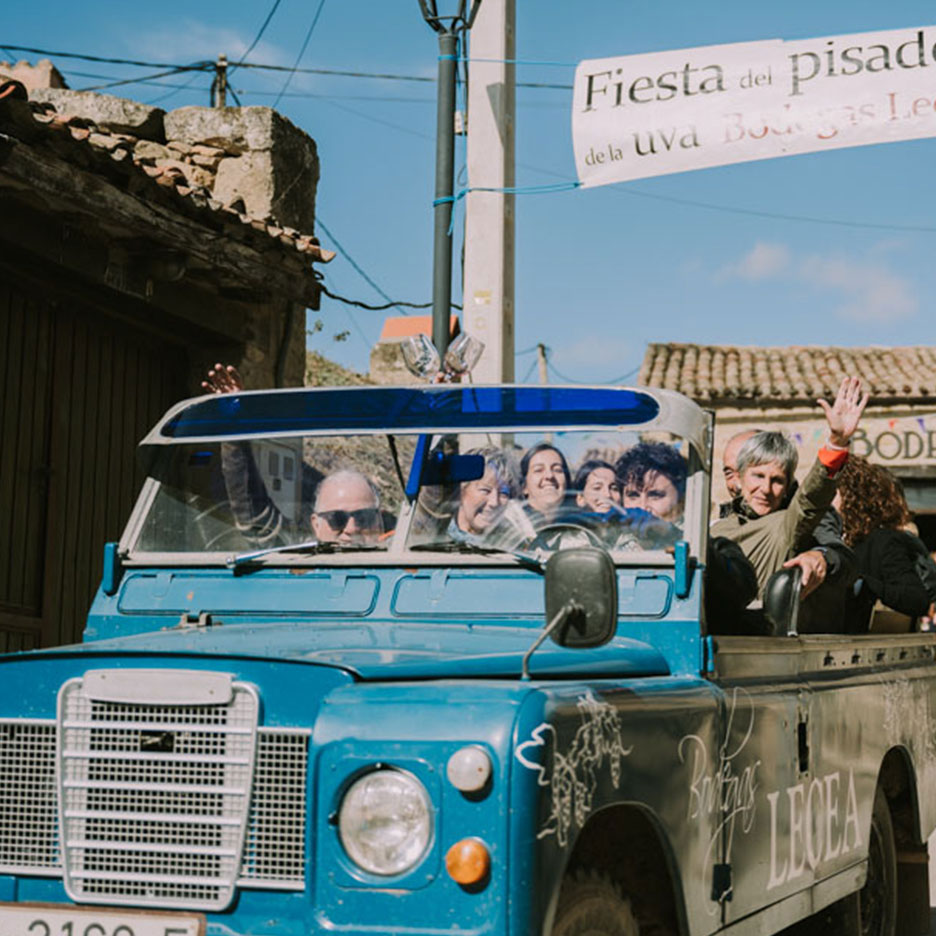Ljudje mahajo iz modrega terenskega vozila med vinskim festivalom, z napisom „Fiesta del pisado de la uva“.