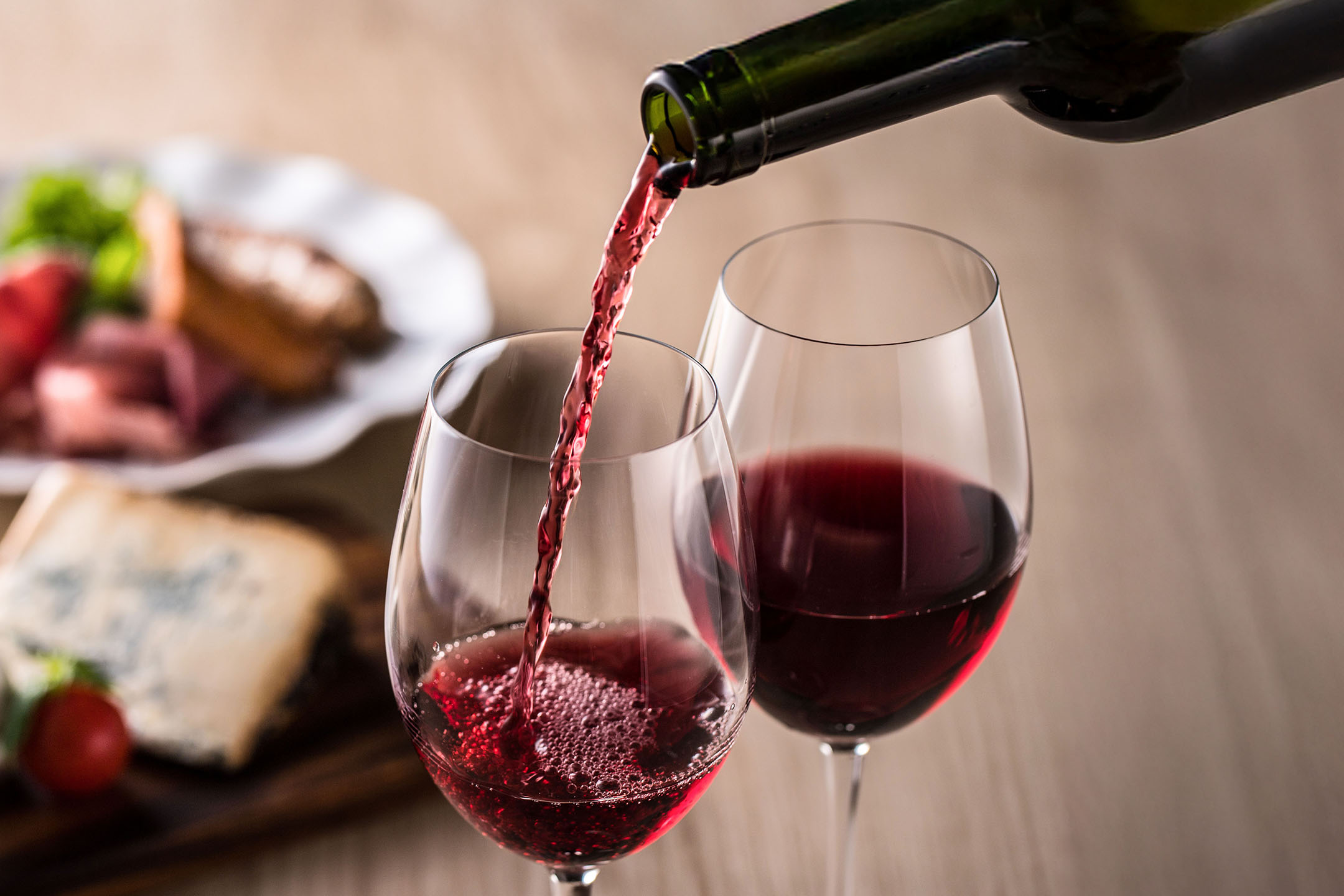 Rdeče vino se toči v kozarec, z predjedmi v ozadju.