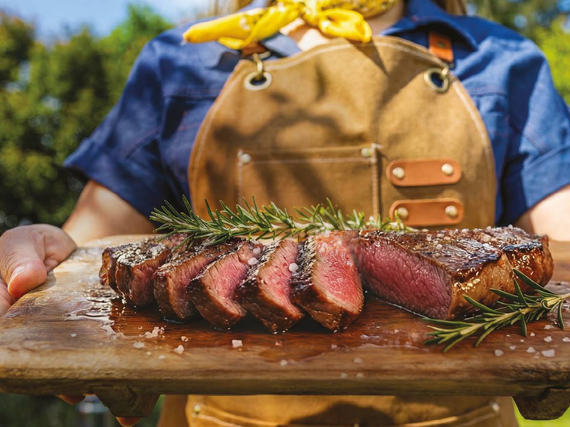 Oseba v predpasniku drži leseno desko z narezanim zrezkom, rožmarinom in soljo.