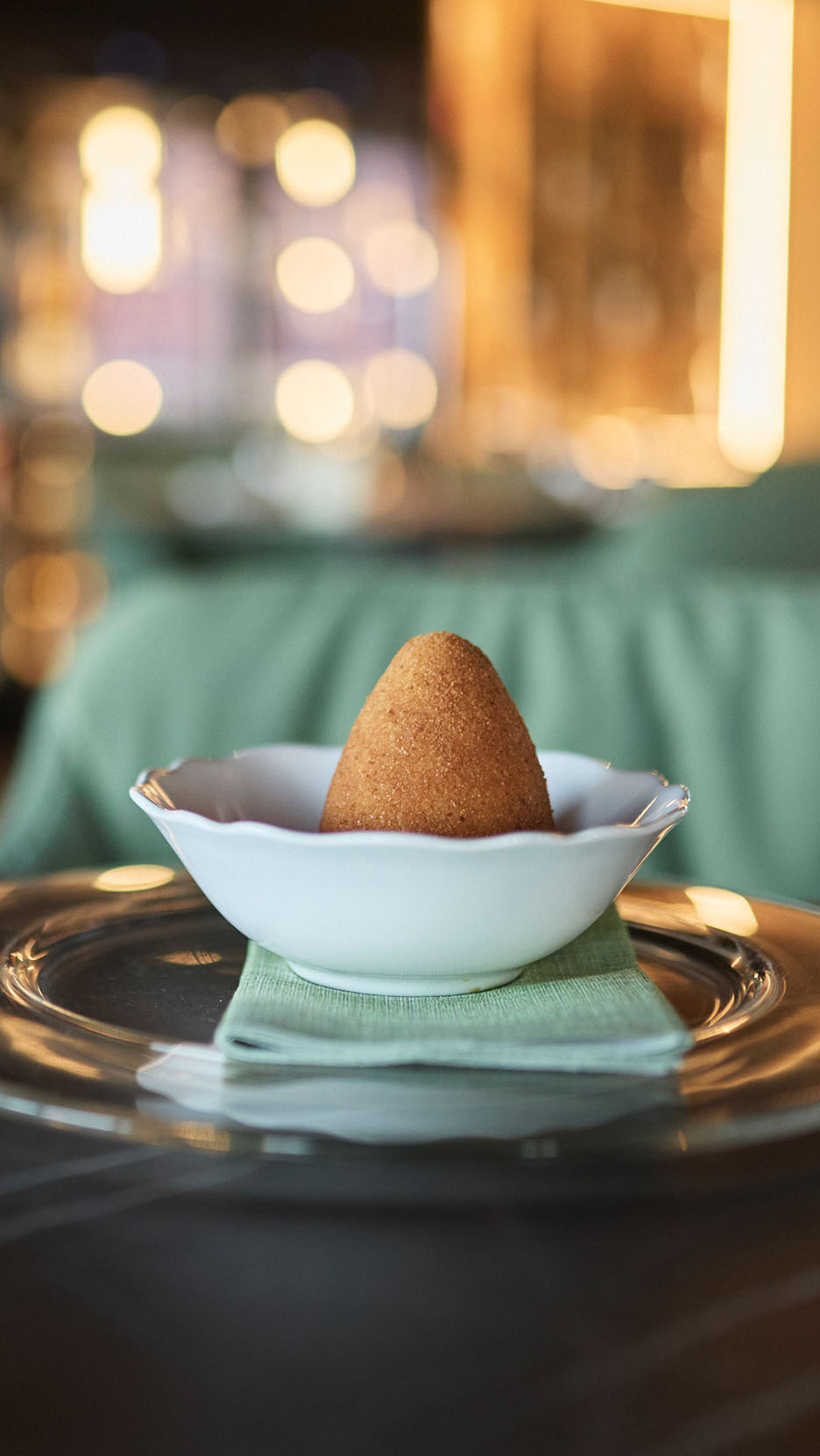 Zlati arancini v beli skledi na zeleni servieti in steklenem krožniku.