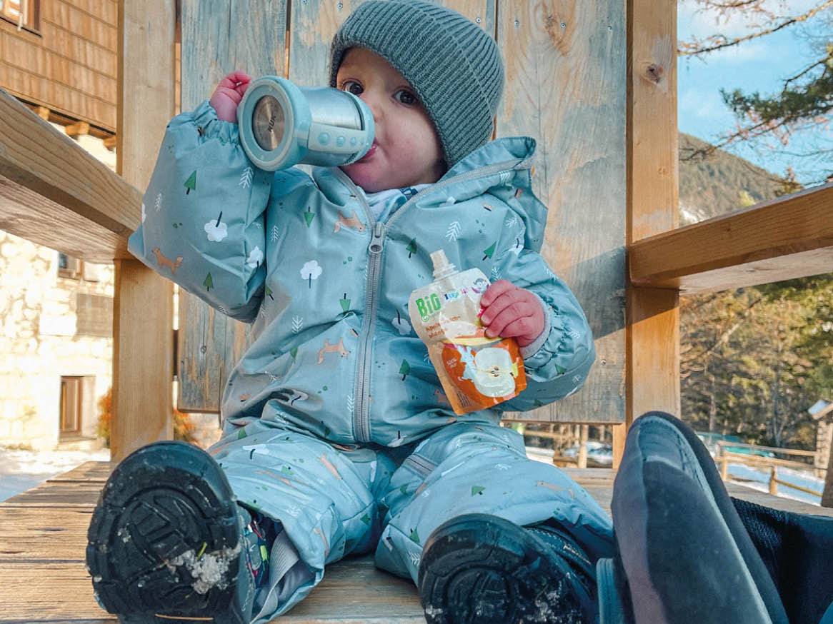 Dojenček v puhastem kombinezonu pije iz termoskopa in uživa sadni pire Bio Happy.