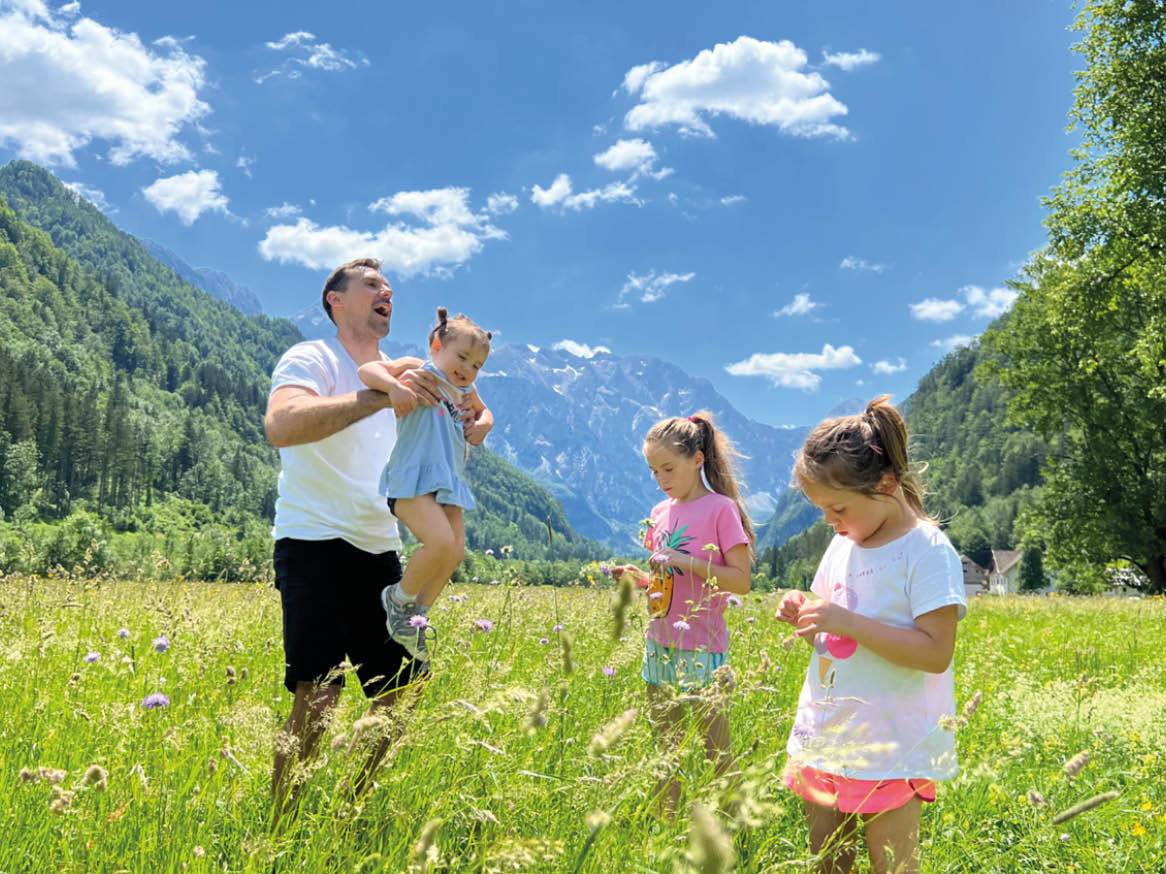 Oče se v zelenem travniku smeji in dvigne svojo hčerko, medtem ko se druge dve hčerki igrajo v bližini.