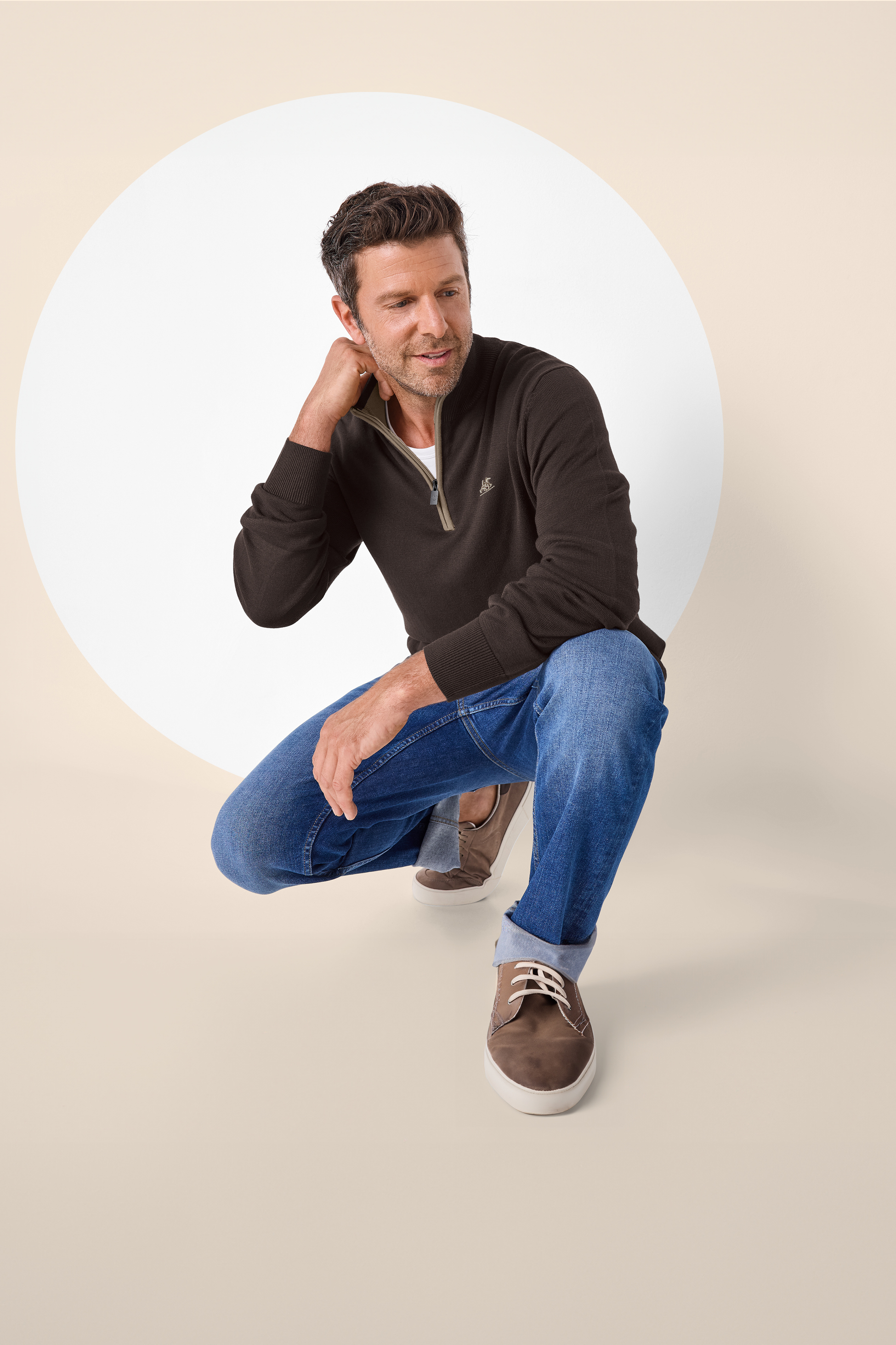 Moški v rjavem puloverju z zadrgo in kavbojkah, čepi pred belim krogom.