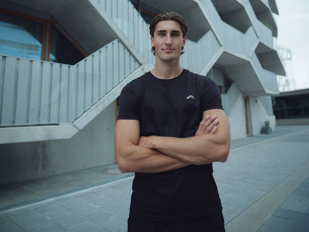 Moški v črni majici z logotipom in črnih kratkih hlačah, stoji zunaj s prekrižanimi rokami.