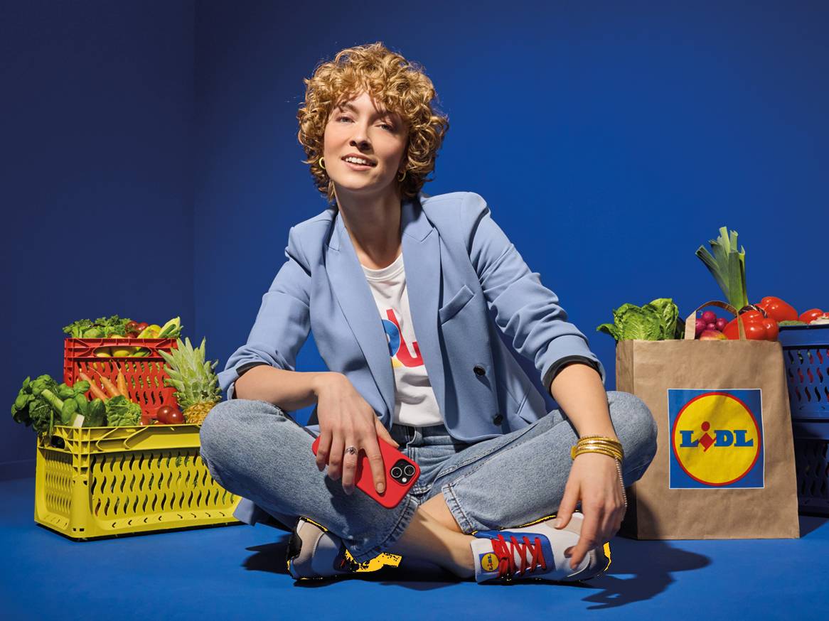 Ženska sedi na tleh, obkrožena s svežo zelenjavo v zabojčkih in nakupovalno vrečko.