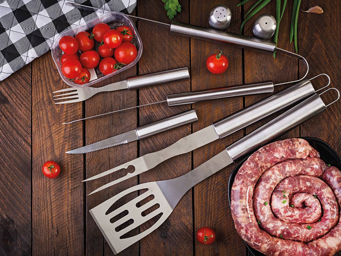 Pripomočki za žar iz nerjavečega jekla, paradižnik in surova klobasa na leseni mizi.