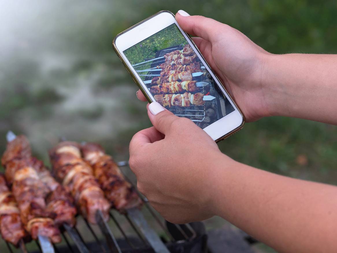 Roke držijo telefon, ki prikazuje fotografijo nabodal na žaru, v ospredju pa so nabodala na žaru.