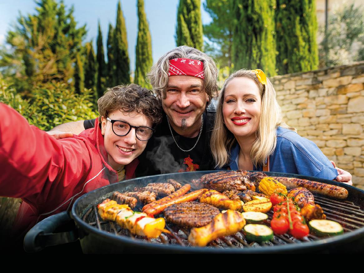 Družina se smehlja ob žaru s piščancem, burgerji, klobasami in zelenjavo.