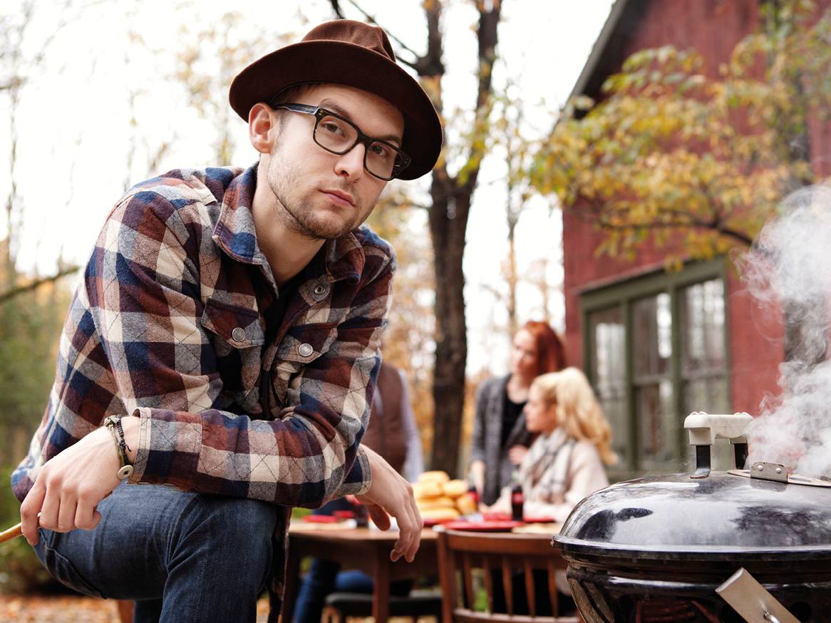Moški v karirasti srajci in klobuku peče na žaru, v ozadju so ljudje in hiša.