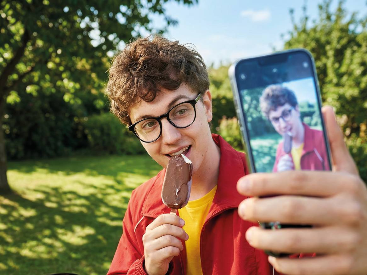 Moški z očali je ugriznil v sladoled na palčki in se slika s telefonom.