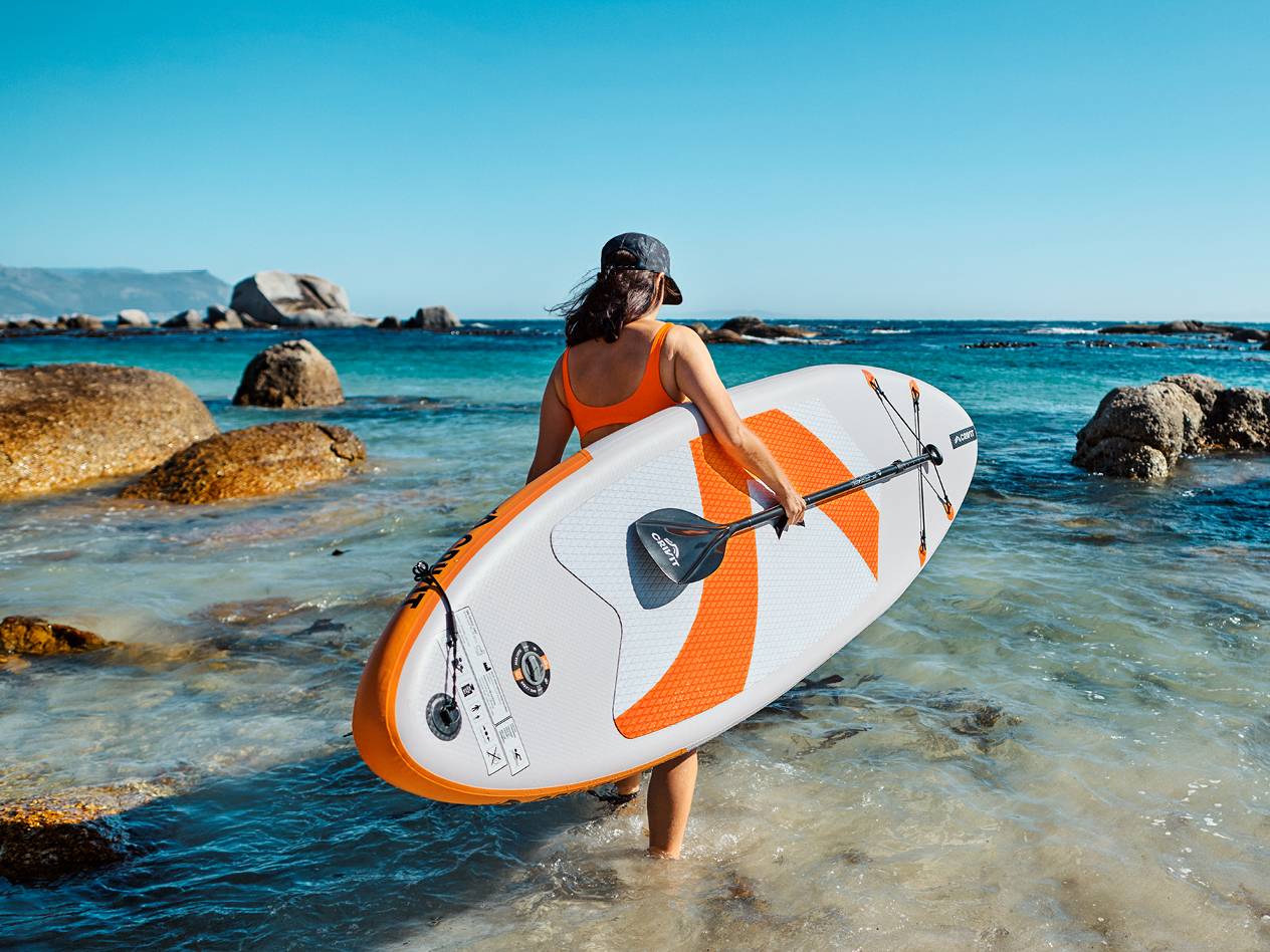 Ženska v kopalkah nosi napihljivo SUP desko Crivit z veslom v plitvi vodi.