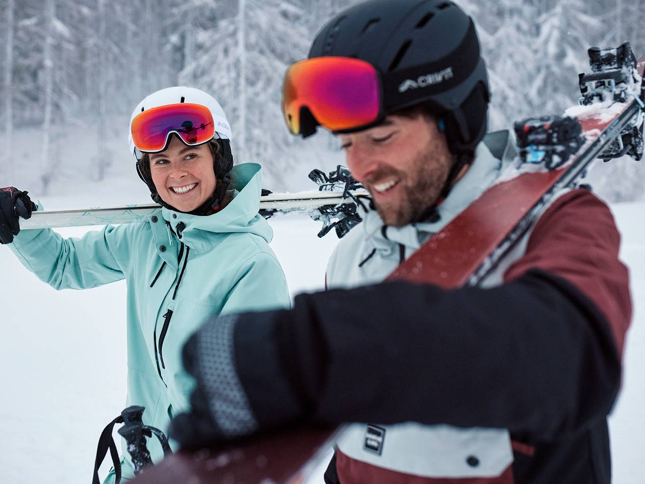 Srečna smučarja v smučarskih oblačilih in čeladah s smučmi na ramenih.