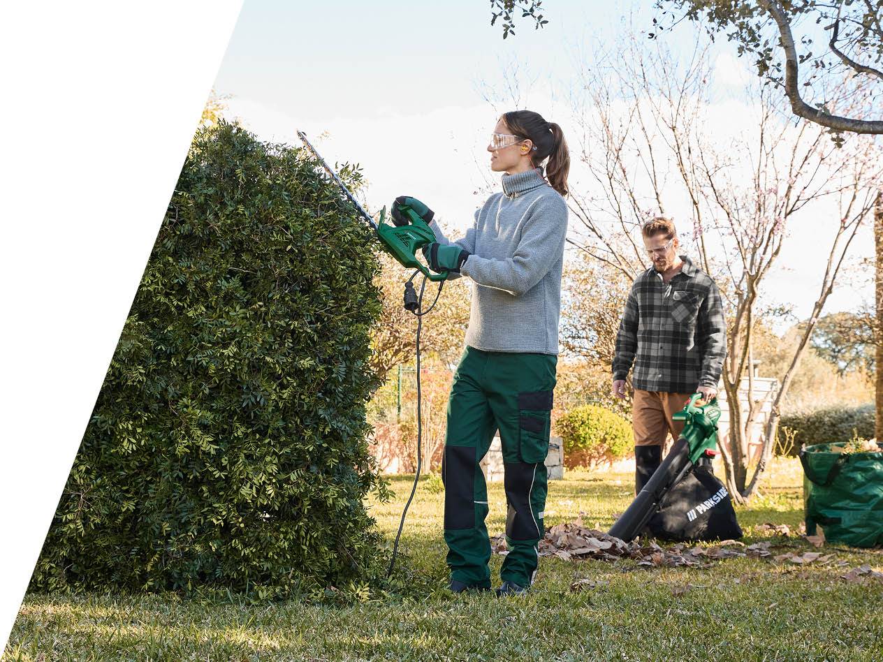 Ženska obrezuje živo mejo s škarjami za živo mejo Parkside, moški pa uporablja pihalnik za listje.