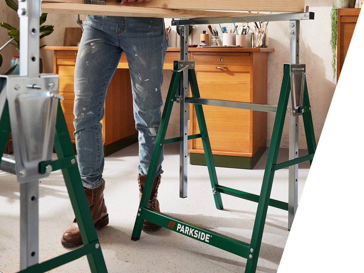 Oseba v delovnih hlačah stoji med dvema zelenima stojaloma Parkside, ki podpirata leseno desko.