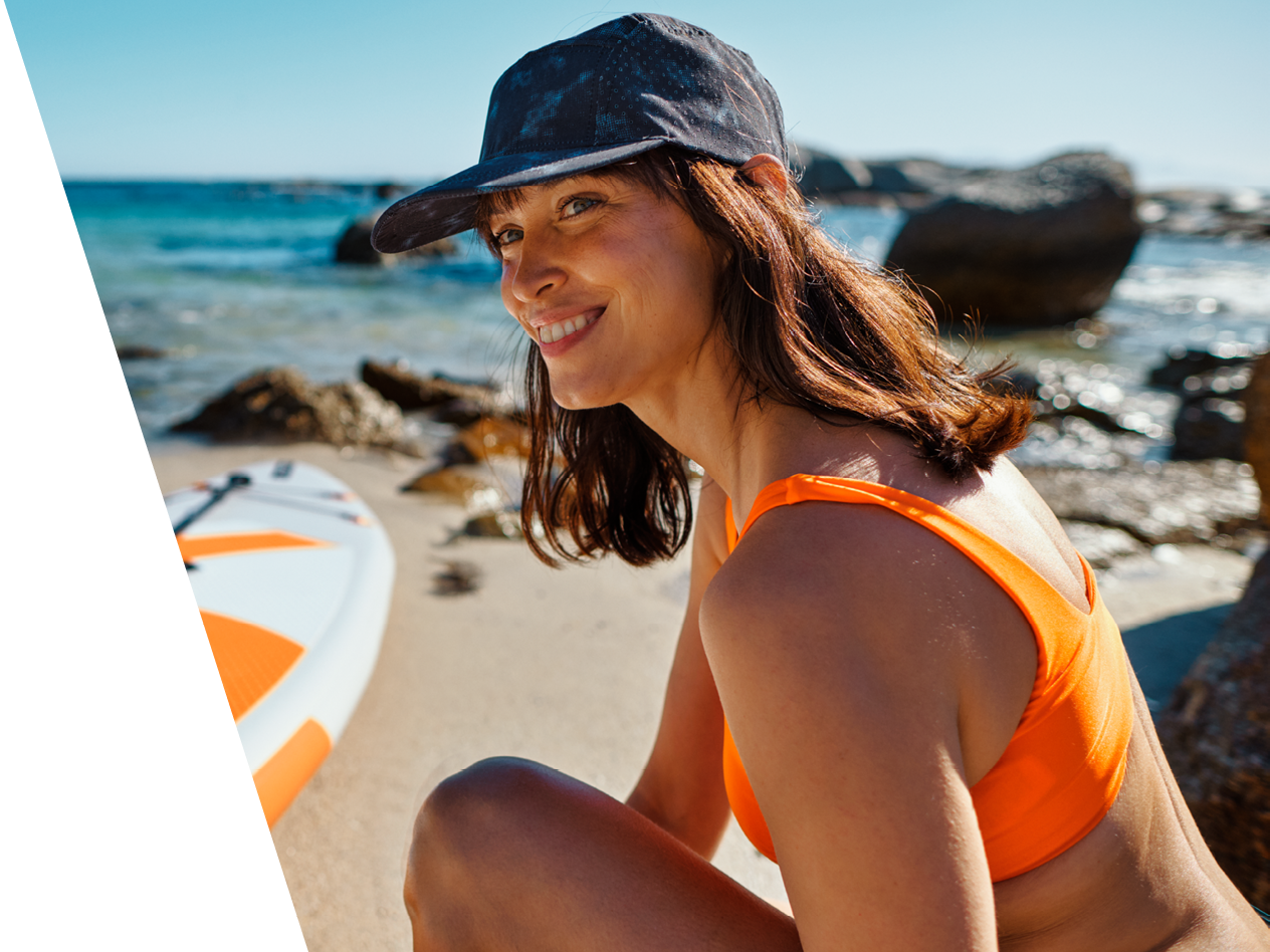 Ženska v oranžnem bikiniju in čepici sedi na peščeni plaži ob deski za SUP.
