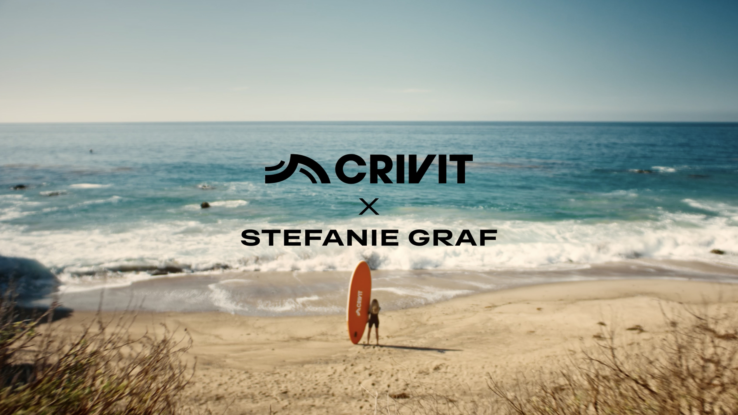 Ženska z oranžno SUP desko na peščeni plaži, z morjem v ozadju.