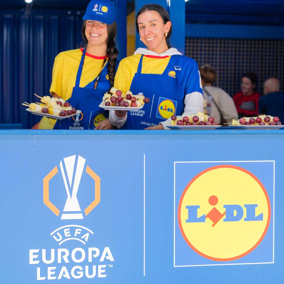 Dve nasmejani ženski v predpasnikih Lidl ponujata sadje na nabodalih na dogodku UEFA Europa League.