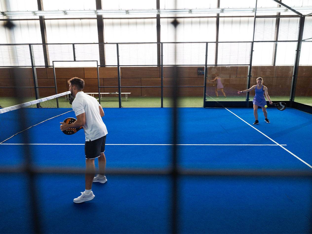 Moški in ženska igrata padel na modrem igrišču z loparji in žogico.