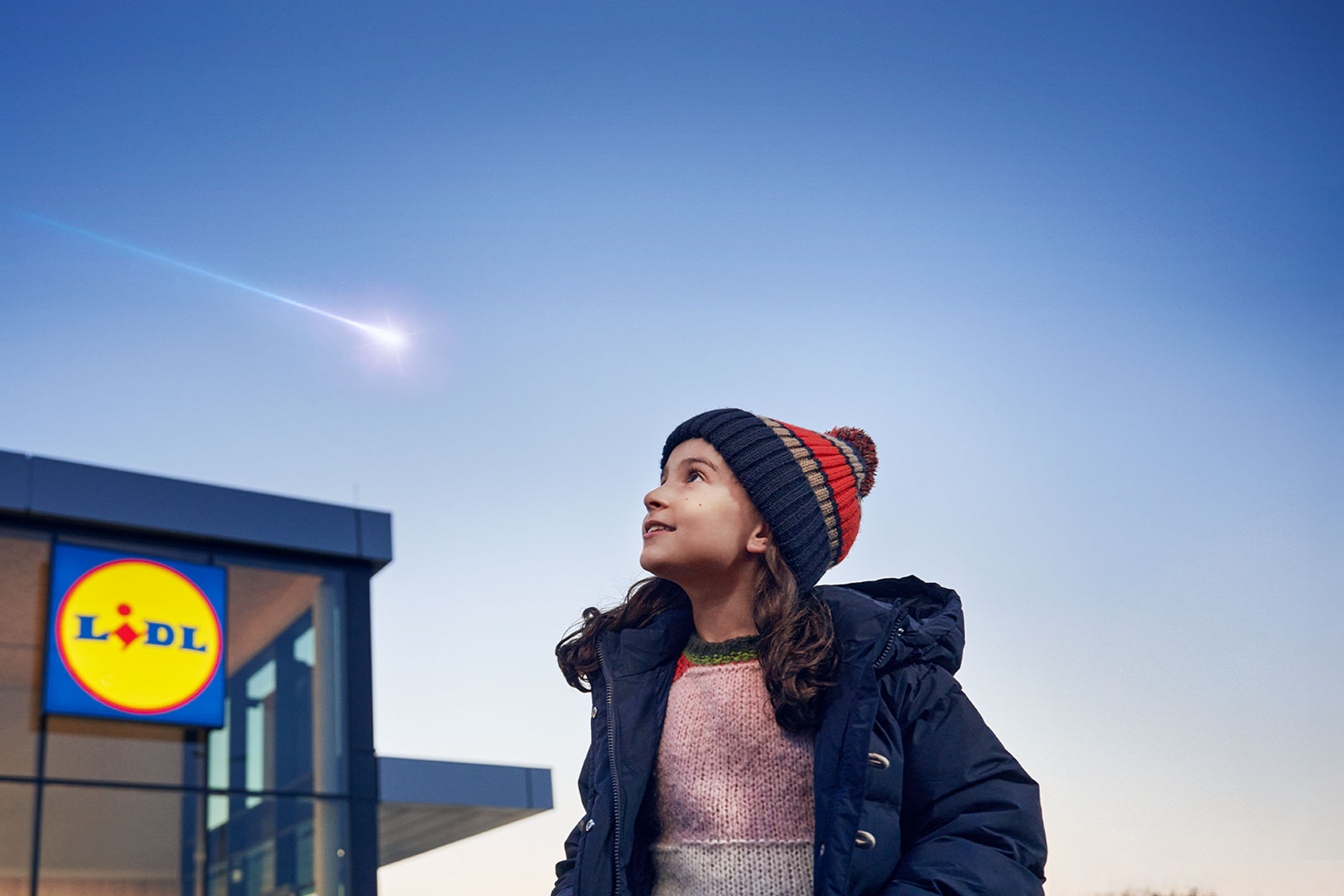 Deklica v kapi gleda meteor na nebu, z trgovino Lidl v ozadju.