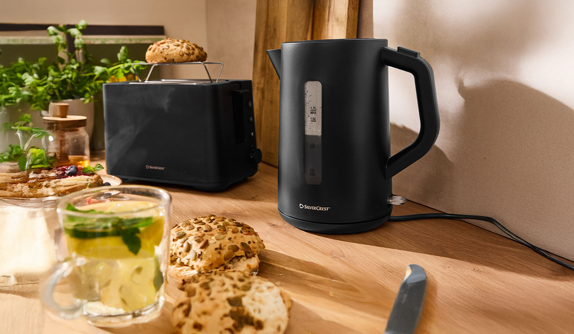 Črn SilverCrest grelnik vode in opekač kruha na leseni mizi z zajtrkom.