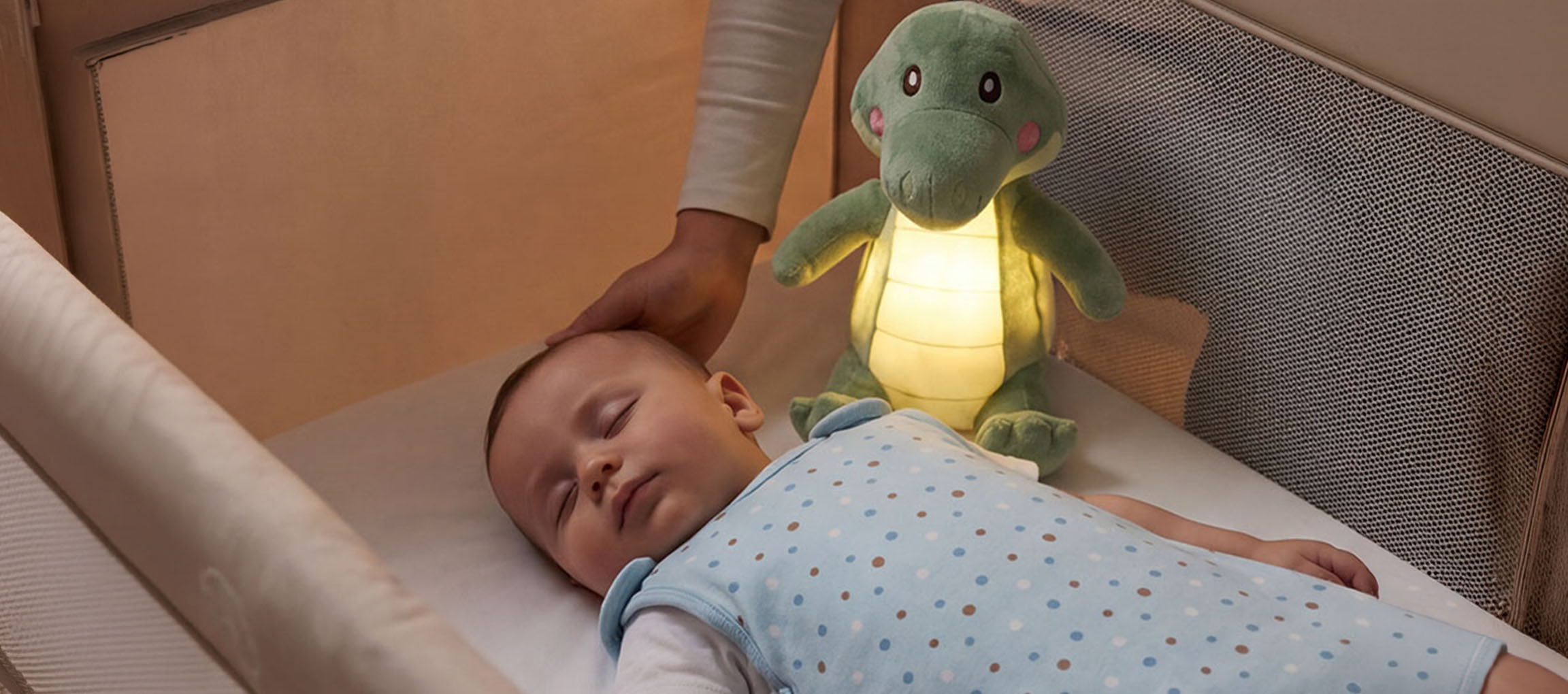 Dojenček spi v posteljici s plišasto nočno lučko, odrasla oseba boža dojenčkovo glavo.