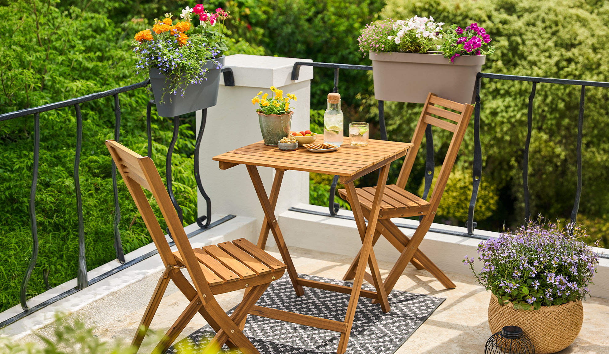 Balkon z lesenim bistro kompletom, lončnicami in pijačami na vzorčasti preprogi.