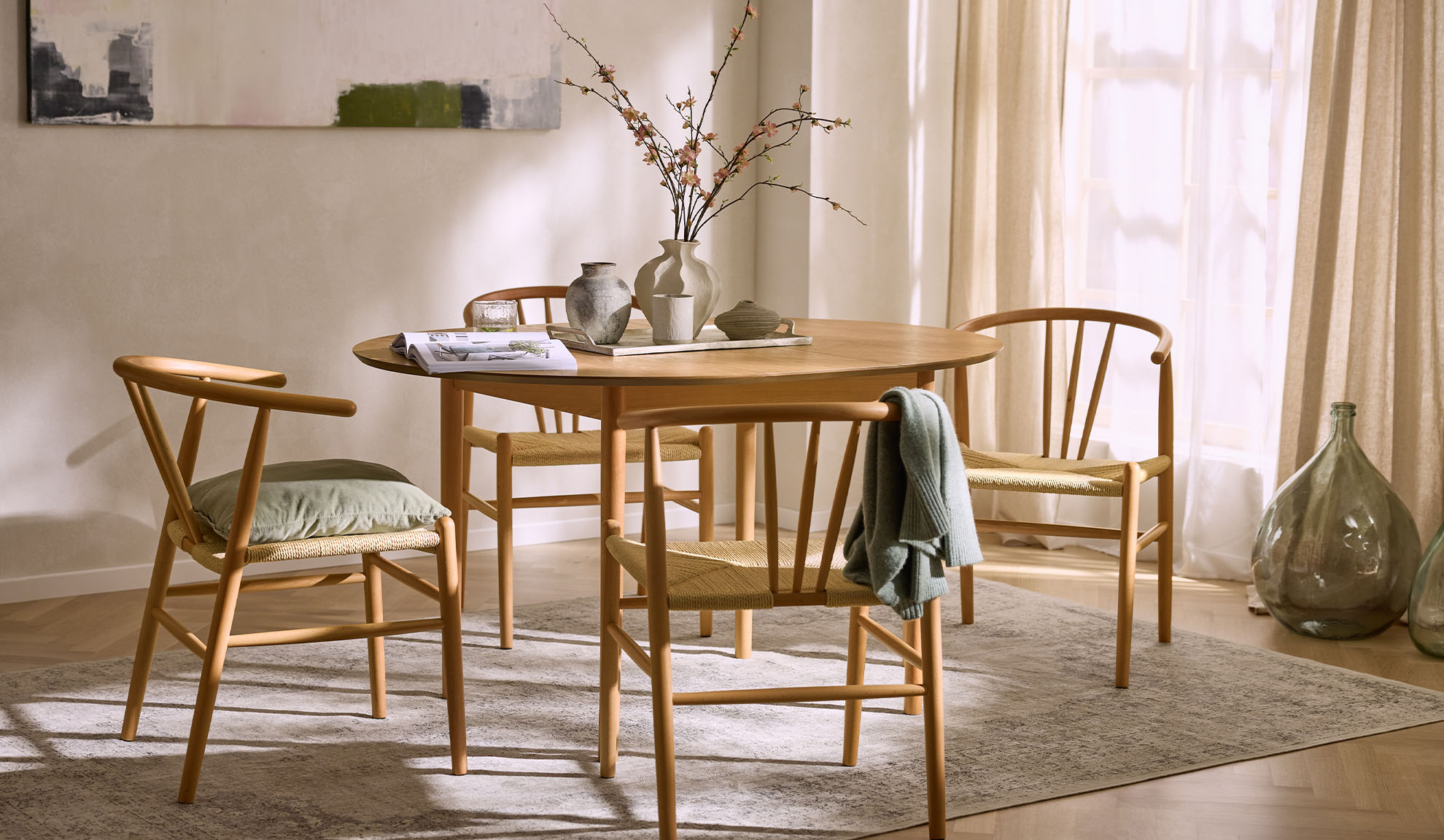 Svetla jedilnica z okroglo leseno mizo, lesenimi stoli z vrvnimi sedeži in cvetlično dekoracijo.