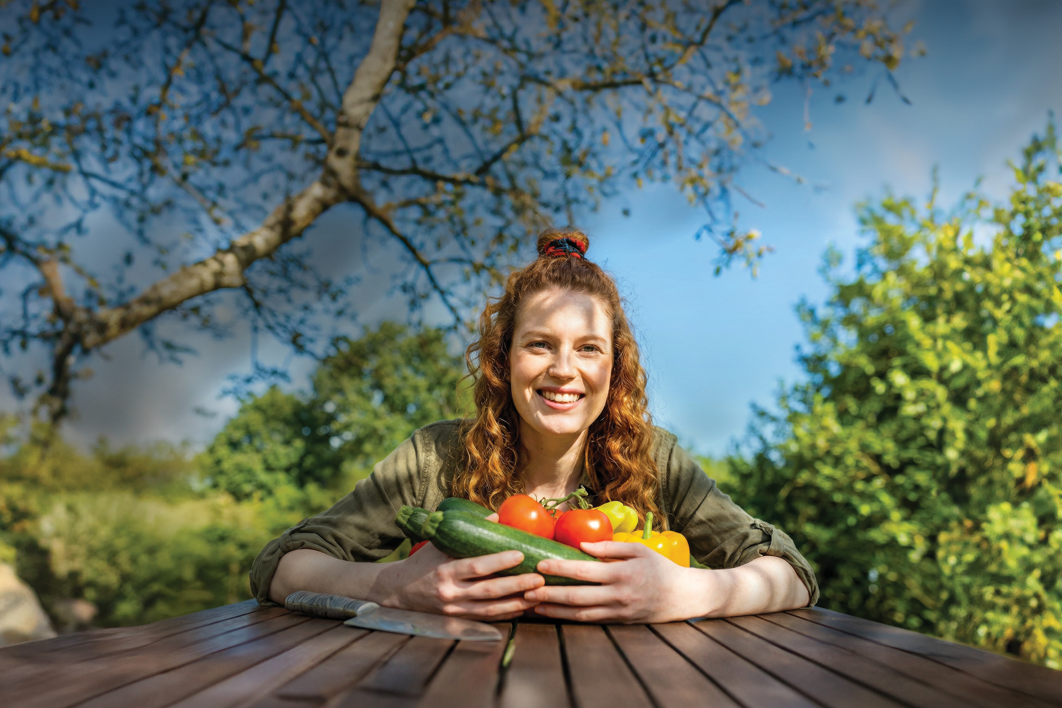 Ženska drži svežo zelenjavo, vključno z bučkami, paradižnikom in papriko, na leseni mizi na prostem.