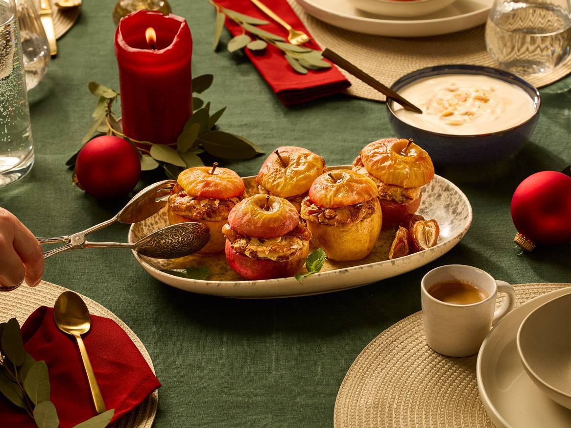 Pekač s pečenimi jabolki, rdeča sveča in božične kroglice na zeleni namizni krpi.