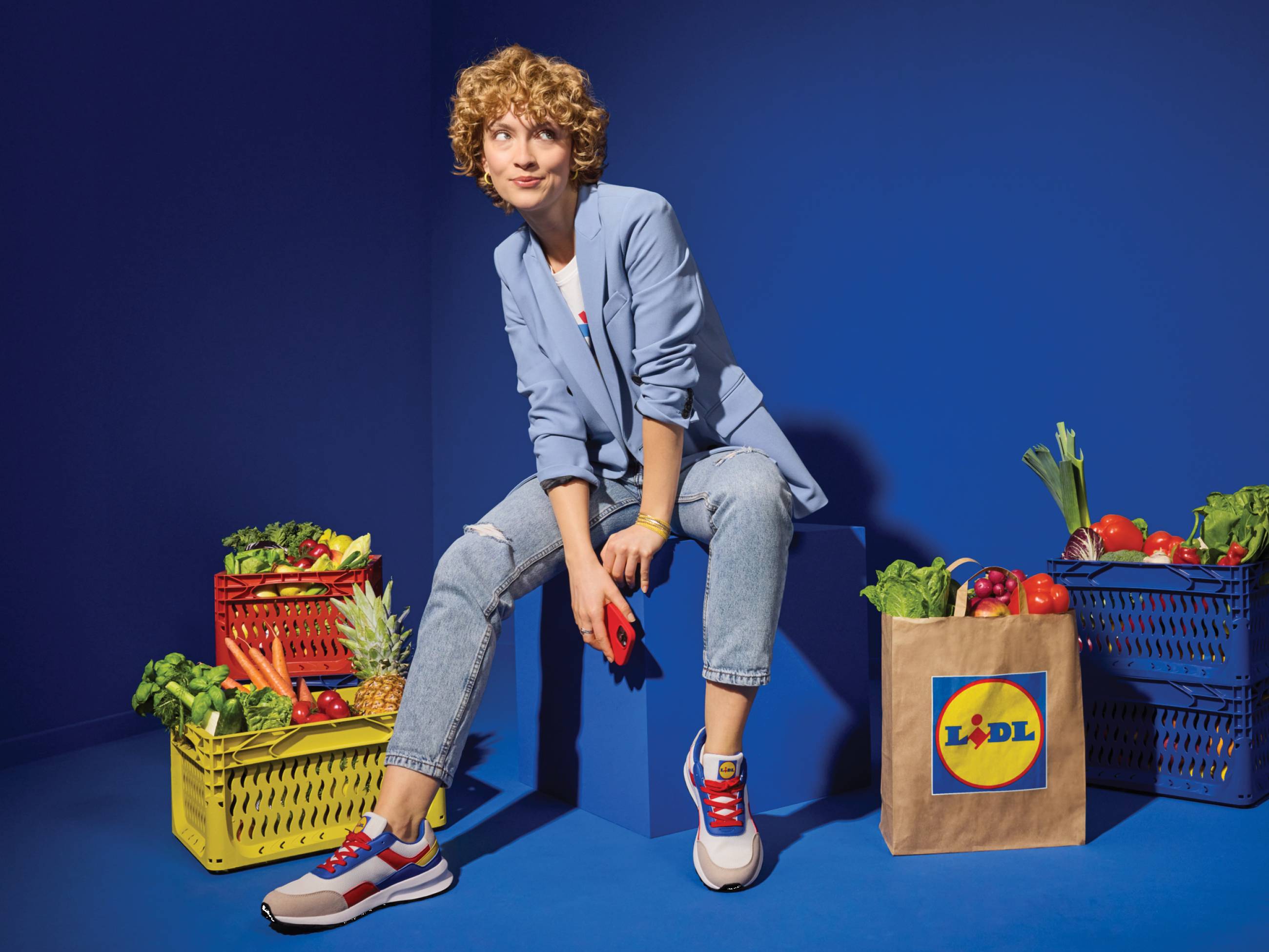 Ženska v modrem suknjiču sedi na modrem bloku z Lidl vrečko in košarami polnimi sadja in zelenjave.