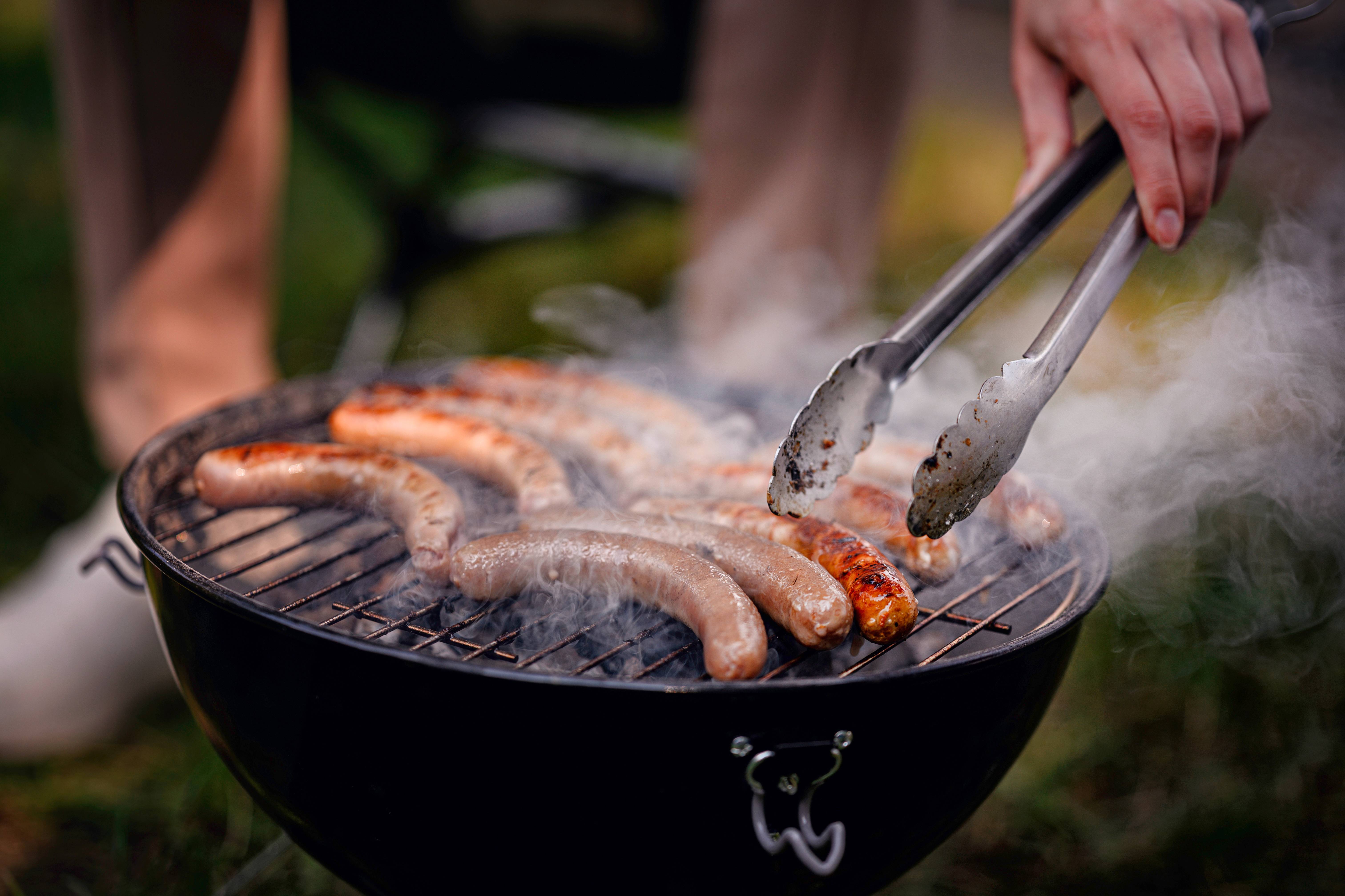 Klobase na žaru, ki jih obrača roka s kleščami, obdane z dimom.