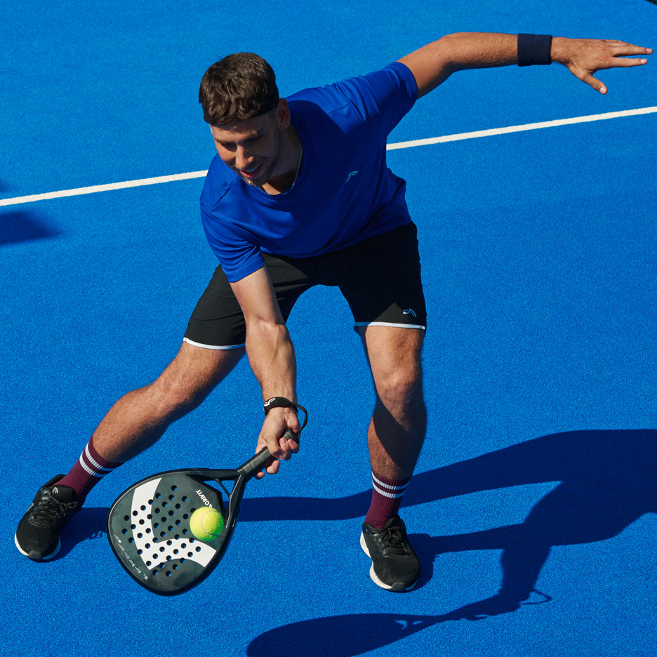 Moški v modri majici in črnih kratkih hlačah igra padel z loparjem in žogico.