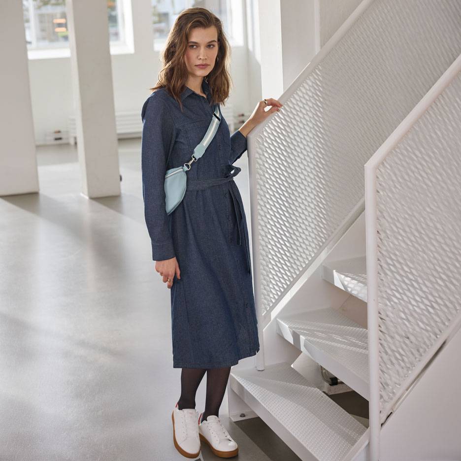 Ženska v jeans srajčni obleki, črnih nogavicah in belih supergah, s svetlo modro torbico okoli pasu.